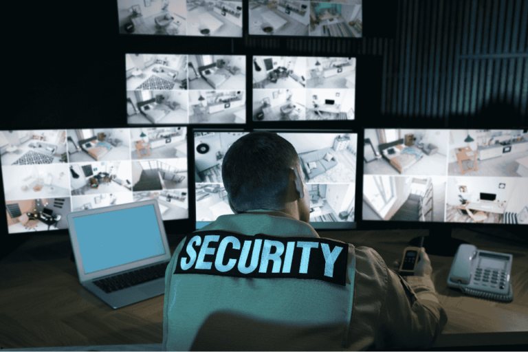 The image shows a security officer sitting at a desk, monitoring multiple surveillance camera feeds displayed on several large screens. The officer wears a jacket labeled "SECURITY" on the back, and the workspace includes a laptop, telephone, and communication device. The scene reflects a control room setup for video surveillance and security monitoring.