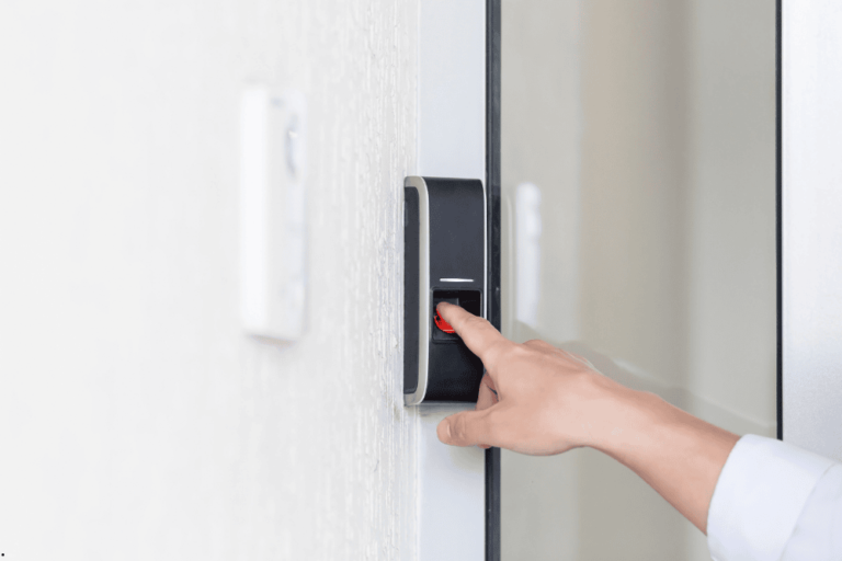 The image shows a person pressing their finger on a biometric fingerprint scanner mounted on a wall near a glass door. The scanner is part of an access control system, allowing entry through fingerprint recognition. The setting suggests a secure environment, such as an office or restricted area, emphasizing modern security technology.