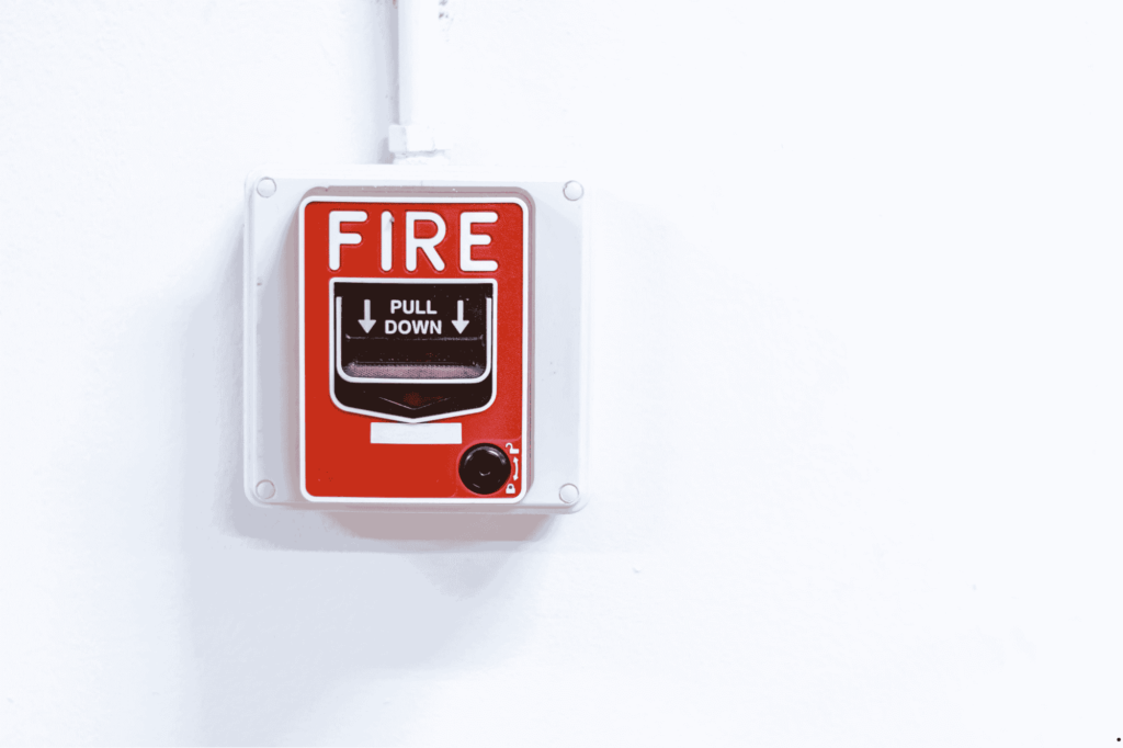 A red fire alarm pull station mounted on a white wall. The alarm has bold white text that reads "FIRE" and instructions below that say "PULL DOWN" with arrows pointing downward.