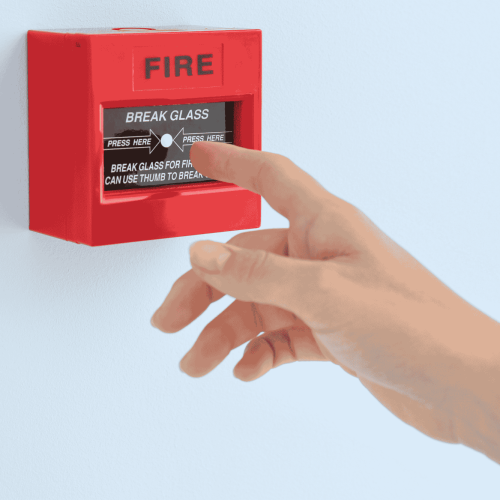 A hand reaching to press a red fire alarm with the words "FIRE" and "BREAK GLASS" on the front. The alarm instructs to "PRESS HERE" and "BREAK GLASS FOR FIRE CAN USE THUMB TO BREAK." The alarm is mounted on a light-colored wall.