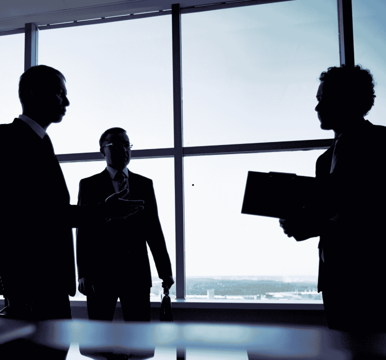 Silhouettes of three business professionals standing in front of large office windows during a meeting, with one holding a folder and the others engaged in conversation. The cityscape is visible in the background through the glass.